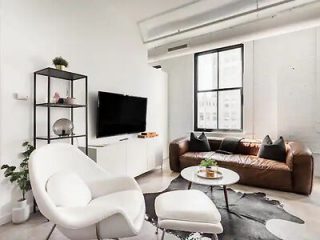 Modern living room in an urban apartment with a brown leather sofa, white armchair, and large windows providing natural light